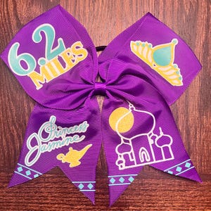 Pode incluir: Laço de cabelo roxo com um design branco e dourado que apresenta uma coroa, uma lâmpada de gênio e uma mesquita. O laço também tem o texto "6.2 Miles" e "Princess Jasmine" nele.