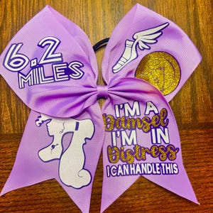 Könnte beinhalten: Lilafarbener Cheer-Bogen mit goldenen Glitzerakzenten. Der Bogen zeigt den Text "6.2 Miles", "I'm a damsel I'm in distress I can handle this" und eine Grafik mit einem geflügelten Fuß.