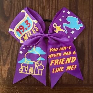 Pode incluir: Um laço de torcida roxo com uma faixa elástica preta. O laço apresenta gráficos e texto do filme Aladdin, incluindo o texto "19.3 Miles" e "You Ain't Never Had A Friend Like Me!"