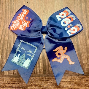 Puede incluir: Un lazo de animadora azul marino con un horizonte de la ciudad de Nueva York en blanco y plata, una silueta de corredor y el texto "1:00:00" en naranja y purpurina plateada.