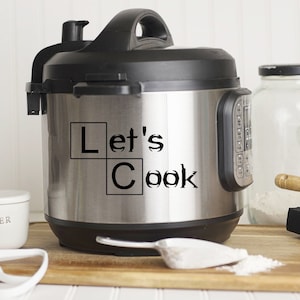 May include: A stainless steel electric pressure cooker with a black lid and a black handle. The cooker has a sticker on the front that says "Let's Cook" in a black and white block font.