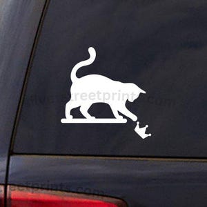 May include: White vinyl decal of a cat reaching for a crown. The cat is on a platform, and the crown is below. The decal is on the rear window of a dark-colored vehicle. The text "silverstreetprints.com" is visible.