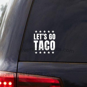 May include: White vinyl decal with the text "LET'S GO TACO" in a bold, sans-serif font, with star accents above and below the text. The decal is affixed to the rear window of a dark-colored vehicle.