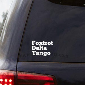 May include: White vinyl decal applied to a car window. The decal features the words "Foxtrot Delta Tango" stacked vertically in a bold, sans-serif font. The website address "silversteeprint.com" is printed below the words.