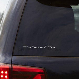 May include: A white vinyl decal featuring Morse code characters applied to the rear window of a dark-colored vehicle. The decal displays a sequence of dots and dashes arranged horizontally, contrasting against the dark glass of the car window.