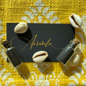 May include: Two small glass bottles with cork tops filled with black sand, four white cowrie shells, and a black card with the word "Aurela" written in gold calligraphy.