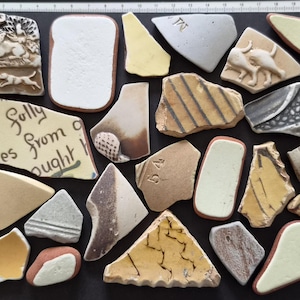 Könnte beinhalten: Eine Sammlung von dreiundzwanzig verschiedenen Keramikscherben in unterschiedlichen Formen, Farben und Texturen, darunter Stücke mit geprägten Tierfiguren, schwarzem Text und Streifenmustern, angeordnet auf einer schwarzen Fläche mit einem 21 cm Lineal.