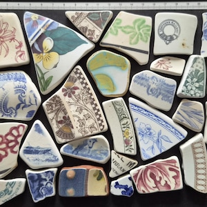 May include: A collection of assorted ceramic shards featuring various patterns, including floral, geometric, and landscape designs in blue, red, green, and yellow. A ruler along the top and left edges shows the pieces range from 1 to 23 cm in size.