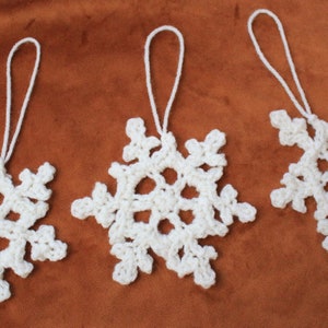 Può includere: Tre decorazioni a forma di fiocco di neve bianche all'uncinetto con anelli per appenderle. I fiocchi di neve sono esposti su uno sfondo marrone. Ogni fiocco di neve ha un design traforato unico, perfetto per le decorazioni natalizie.