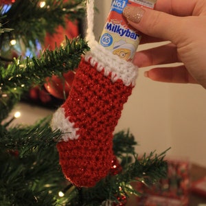 Può includere: Un calzino di Natale rosso e bianco all'uncinetto con una barretta di cioccolato Milkybar al suo interno. Il calzino è appeso a un albero di Natale con luci bianche.