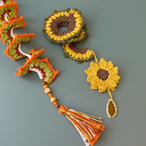 May include: A crocheted sunflower garland with brown and green yarn, accented with wooden beads and a tassel. The garland is made up of multiple sunflower shapes, with a larger sunflower at the end.