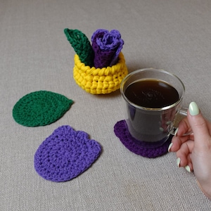 May include: A set of crocheted coasters in purple and green, with a crocheted basket filled with purple and green yarn. A glass mug with coffee sits on a purple coaster.