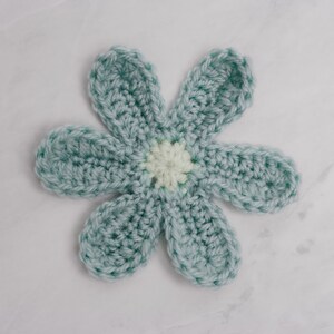 May include: A crocheted flower with six petals in a light blue color. The center of the flower is a white circle with a light yellow center.