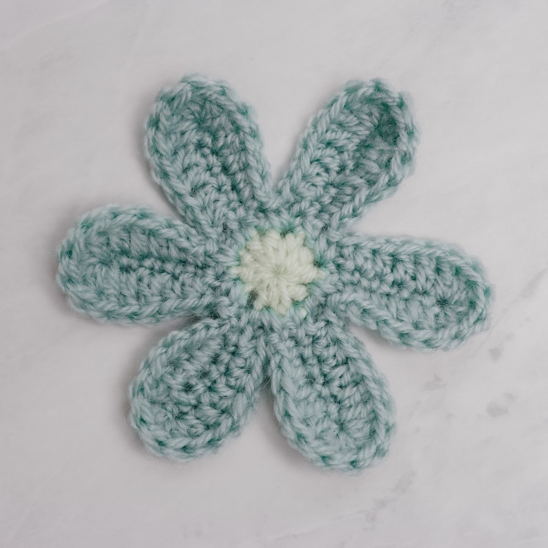 May include: A crocheted flower with six petals in a light blue color. The center of the flower is a white circle with a light yellow center.