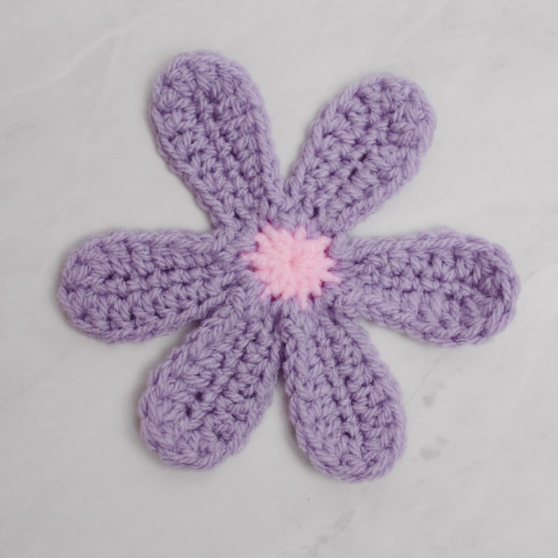 May include: A crocheted flower with six petals in a light purple color. The center of the flower is a pink circle.