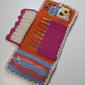 Puede incluir: Estuche de ganchillo naranja con un bolsillo con cremallera rosa y un soporte para ganchos rosa. El estuche tiene un borde festoneado crema y contiene varios ganchillos. Se ven detalles decorativos de flores y alfileres de ganchillo.