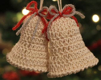 Puede incluir: Dos campanas blancas de ganchillo con lazos de cinta roja y plateada colgando de un árbol de Navidad.