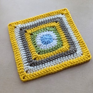 Può includere: Un quadrato all'uncinetto con un design multicolore. Il centro del quadrato è un cerchio blu chiaro circondato da verde, bianco e grigio. Il bordo esterno del quadrato è giallo.