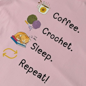 Può includere: Felpa rosa chiaro con le parole "Coffee. Crochet. Sleep. Repeat!" in nero. Il design include illustrazioni di una tazza di caffè, gomitoli di lana, un uncinetto, un gatto che dorme e un simbolo di freccia ripetuta.