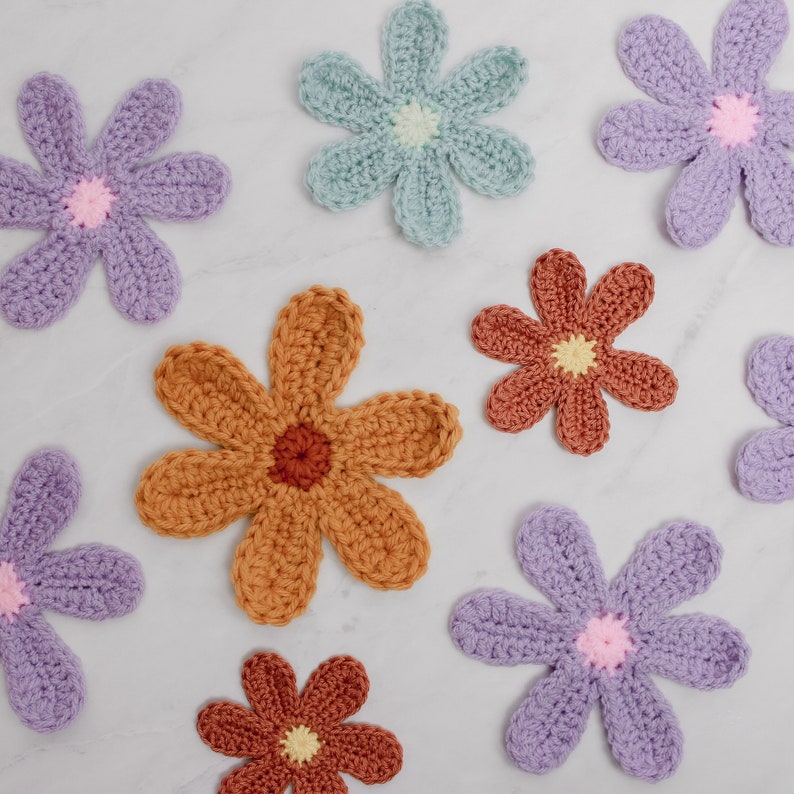 May include: A collection of seven crocheted flower appliques in various colors, including purple, blue, orange, and brown. The flowers have a simple, daisy-like design with multiple petals.