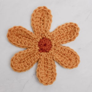 May include: A crocheted flower with six petals in a mustard yellow color. The center of the flower is a smaller circle in a burnt orange color.