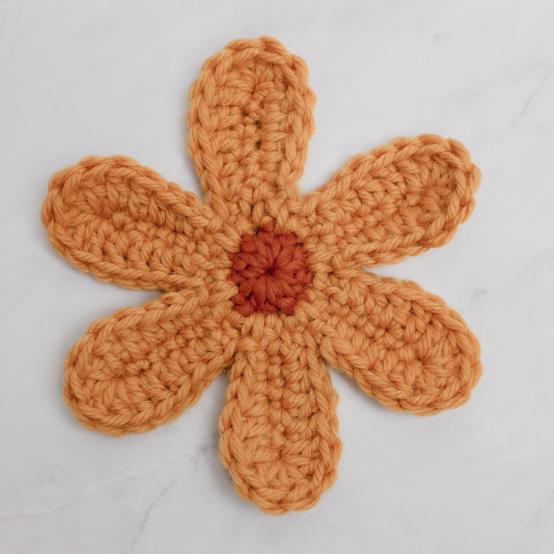 May include: A crocheted flower with six petals in a mustard yellow color. The center of the flower is a smaller circle in a burnt orange color.