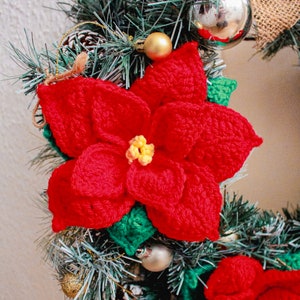Op de afbeelding: Een rode gehaakte kerstster met groene bladeren is bevestigd aan een kerstkrans. De bloem is een populaire kerstdecoratie.