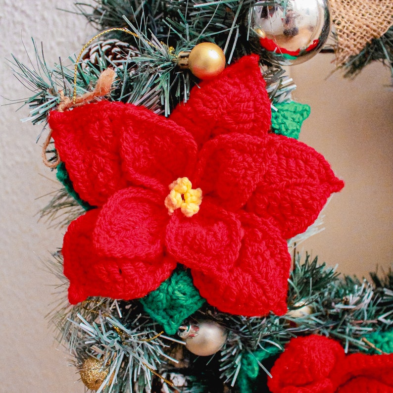 Op de afbeelding: Een rode gehaakte kerstster met groene bladeren is bevestigd aan een kerstkrans. De bloem is een populaire kerstdecoratie.