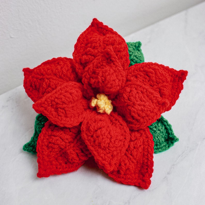Op de afbeelding: Een gehaakte kerststerbloem met rode bloemblaadjes en groene bladeren. De bloem is gemaakt van garen en heeft een gele kern.