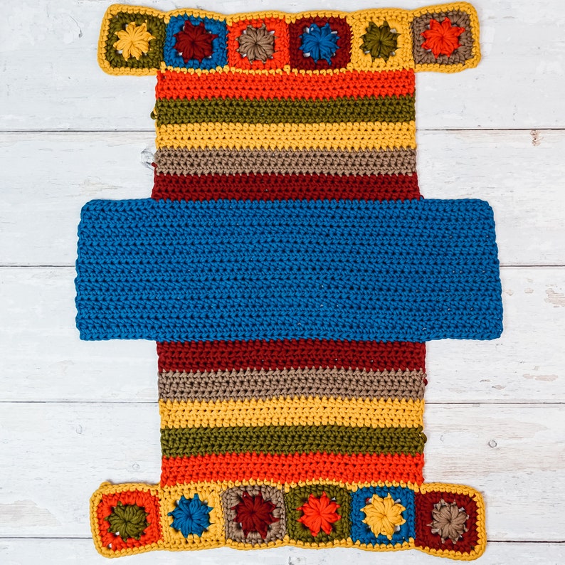 Puede incluir: Un tapiz de pared de crochet con una secci&oacute;n central azul y un borde rayado multicolor. El borde est&aacute; decorado con flores de crochet de colores.