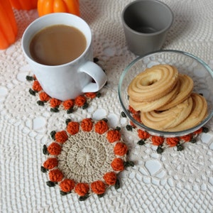 May include: Three crocheted coasters with an orange pumpkin design. The coasters are on a white crocheted tablecloth with a cup of coffee and a bowl of biscuits.