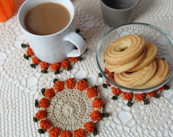 May include: Three crocheted coasters with an orange pumpkin design. The coasters are on a white crocheted tablecloth with a cup of coffee and a bowl of cookies.