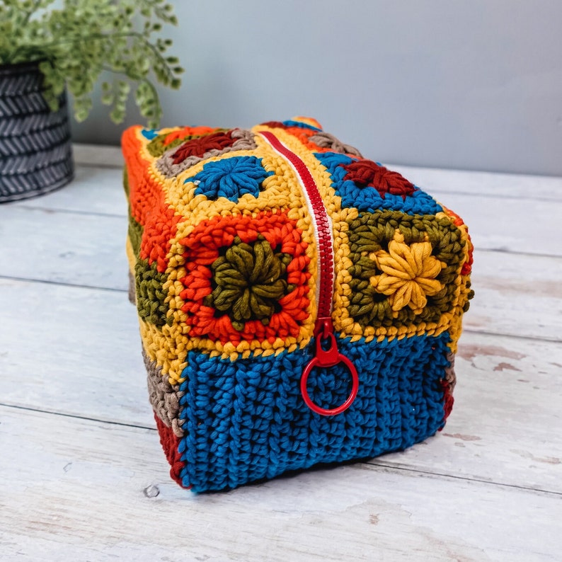 Puede incluir: Una bolsa de cremallera de ganchillo de colores con un tirador de cremallera rojo. La bolsa est&aacute; hecha de cuadrados de diferentes colores en tonos de azul, verde, amarillo, naranja y rojo.