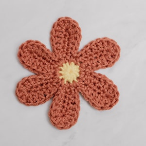 May include: A crocheted flower with six petals in a terracotta color. The center of the flower is a yellow crocheted circle.