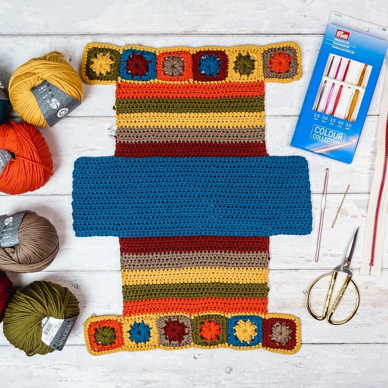 Puede incluir: Una bolsa de ganchillo con un forro azul y rayas de colores. La bolsa est&aacute; hecha con una variedad de colores, incluyendo rojo, naranja, amarillo, verde y azul. La bolsa est&aacute; sentada sobre una superficie blanca con ovillos de lana y ganchillos.