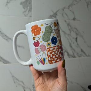 Può includere: Tazza in ceramica bianca con un design colorato che presenta filato, uncinetti e un quadrato all'uncinetto. Il design è in tonalità di rosa, blu, verde, arancione e giallo.