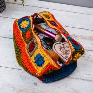 Puede incluir: Una bolsa de cremallera de ganchillo con un patr&oacute;n de rayas de colores y motivos florales. La bolsa est&aacute; abierta y llena de varios art&iacute;culos de maquillaje, incluyendo un pincel de maquillaje rosa, un pincel de maquillaje de madera y una peque&ntilde;a caja en forma de coraz&oacute;n que dice "Diosa del amor".