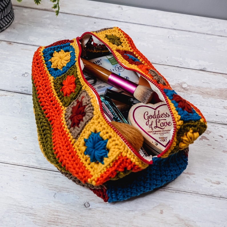 Puede incluir: Una bolsa de cremallera de ganchillo con un patr&oacute;n de rayas de colores y motivos florales. La bolsa est&aacute; abierta y llena de varios art&iacute;culos de maquillaje, incluyendo un pincel de maquillaje rosa, un pincel de maquillaje de madera y una peque&ntilde;a caja en forma de coraz&oacute;n que dice "Diosa del amor".