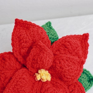 Op de afbeelding: Een gehaakte kerststerbloem met rode bloemblaadjes en groene bladeren. Het midden van de bloem is geel.