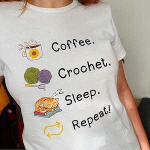 Può includere: T-shirt bianca con un design grafico raffigurante una tazza di caffè, gomitoli di lana, un gatto che dorme e le parole "Coffee. Crochet. Sleep. Repeat!" in nero. La tazza di caffè ha un design a girasole. Il gatto è arancione e bianco.