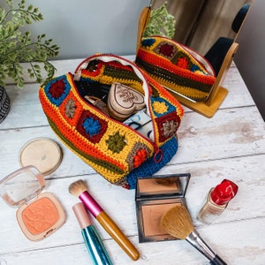 Puede incluir: Una bolsa de maquillaje de ganchillo con un patr&oacute;n geom&eacute;trico colorido. La bolsa est&aacute; abierta y llena de pinceles de maquillaje, un compacto y un jab&oacute;n en forma de coraz&oacute;n. La bolsa est&aacute; sobre una superficie de madera blanca.