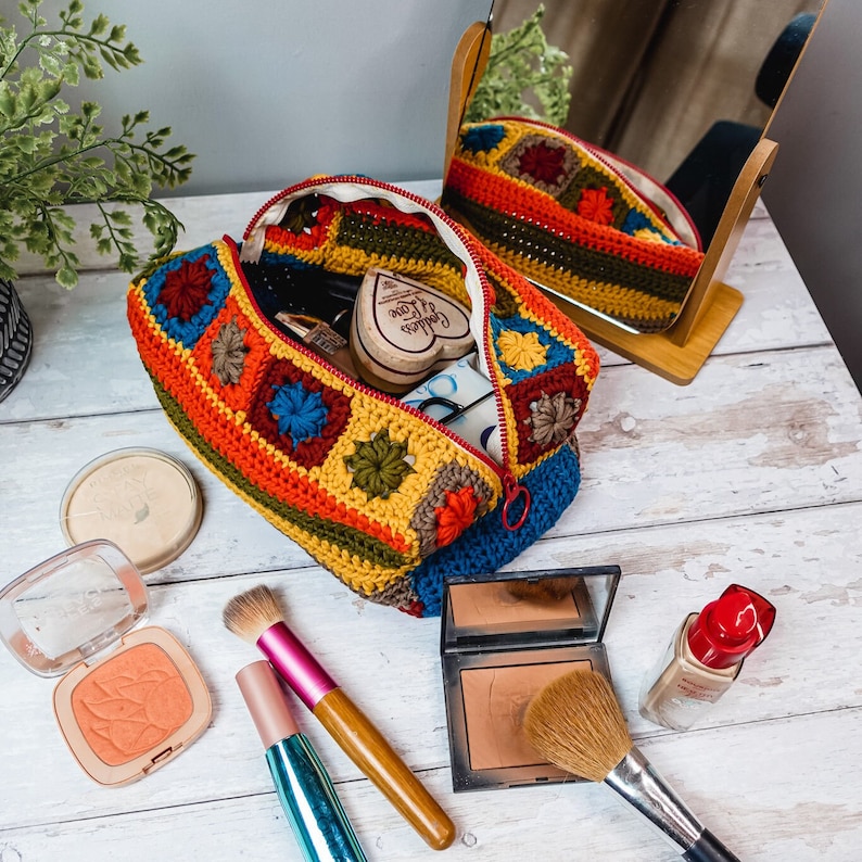 Puede incluir: Una bolsa de maquillaje de ganchillo con un patr&oacute;n geom&eacute;trico colorido. La bolsa est&aacute; abierta y llena de pinceles de maquillaje, un compacto y un jab&oacute;n en forma de coraz&oacute;n. La bolsa est&aacute; sobre una superficie de madera blanca.