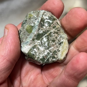 Peut inclure: Une pierre lisse, verte et blanche avec un motif marbré. La pierre a une forme légèrement arrondie et fait environ la taille d'une balle de golf.