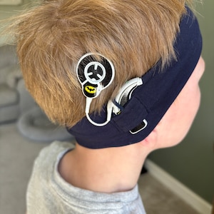 Könnte beinhalten: Ein junges Kind trägt ein blaues Stirnband mit einem schwarzen und gelben Batman-Logo. Ein Hörgerät ist am Stirnband befestigt und ein kleines rundes Gerät mit einem schwarz-weißen Design ist am Stirnband befestigt.