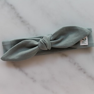 May include: A sage green fabric headband with a knotted bow. The headband has a small white label with the letters "MC" on it.