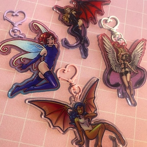 May include: Collection of fantasy-themed keychains featuring illustrations of winged figures, including fairies and demons. The keychains have various colors, including red, blue, and purple, with heart-shaped clasps and a pink background.