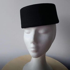 May include: A black, rectangular hat with a flat top and a slightly curved brim. The hat is made of a textured fabric and is displayed on a white mannequin head. The hat is a classic style, suitable for various occasions.