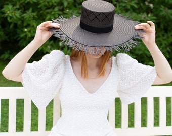 Puede incluir: Una mujer con un vestido blanco y un sombrero negro de ala ancha con ribete de flecos. El sombrero tiene una cinta negra alrededor de la corona.