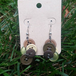 May include: A pair of dangle earrings made with brown, green, and beige buttons. The earrings are hanging from a silver hook.