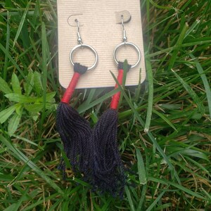 May include: A pair of silver hoop earrings with black and red tassel pendants. The earrings are displayed on a brown card.